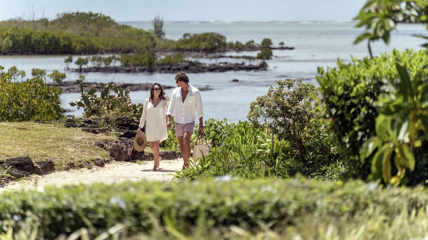 Couple holds hands as they walk along sand path, lagoon in the background