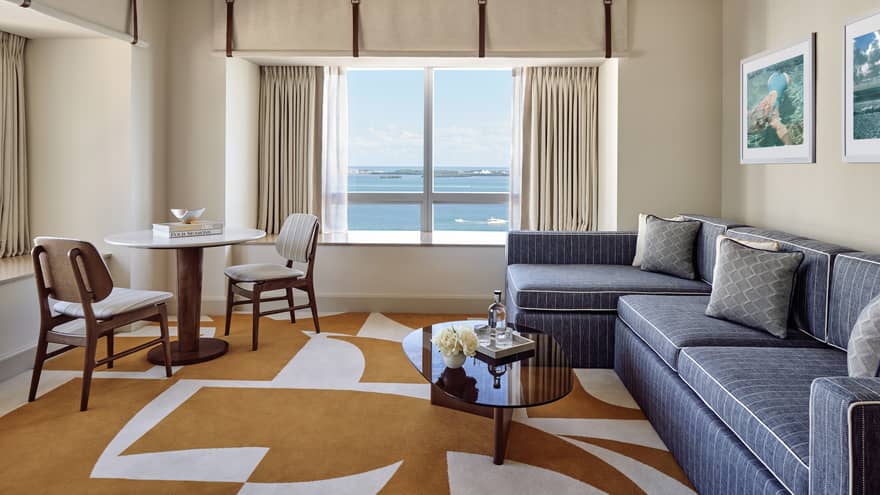 Hotel suite living room with denim-coloured sofa, orange and white rug, table and two chairs, and a window with bay view