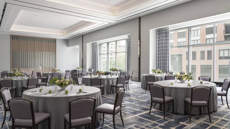 A ballroom with round tables covered in grey table cloth and yellow flower, with large windows in the room.