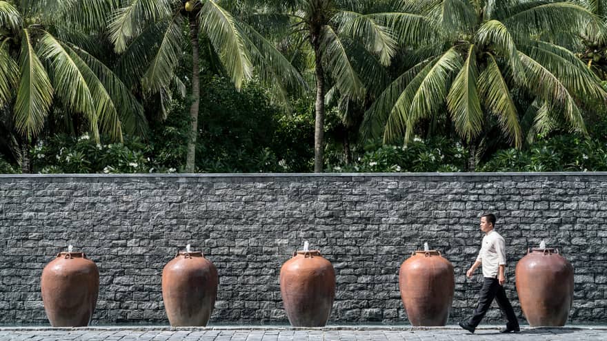 Man walks by palm trees, tall stone fence lined with large clay pot fountains