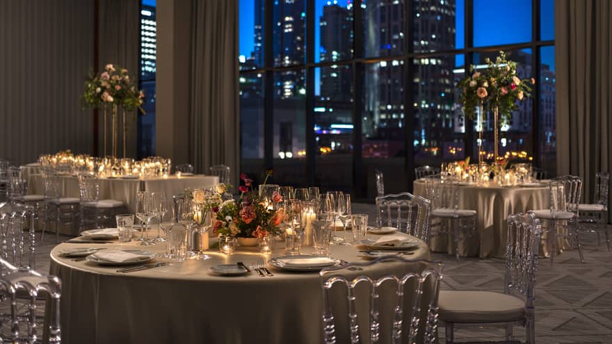 Elegantly set dining tables with floral centerpieces and candles in a dimly lit ballroom, overlooking a city skyline at night.