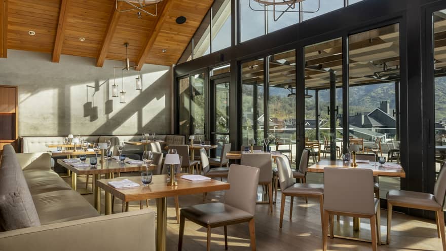 Modern restaurant interior with high wooden ceilings, large windows, and natural light. Tables are set, and an outdoor terrace offers views of surrounding hills.