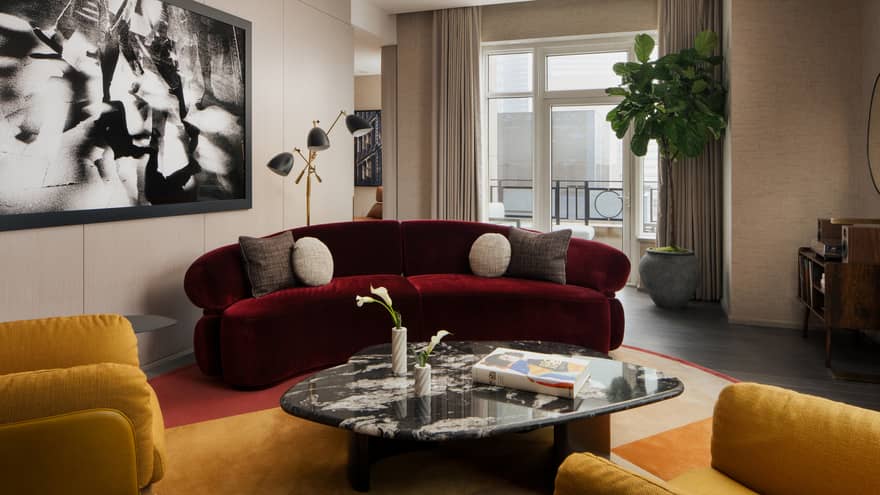 Luxury suite living room with burgundy sofa, coffee table, two yellow armchairs, large black-and-white wall art, potted plant and city-view balcony