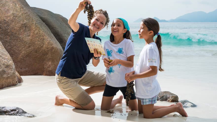 Woman kneels on white sand beach beside boulder, holds up seaweed to show two girls