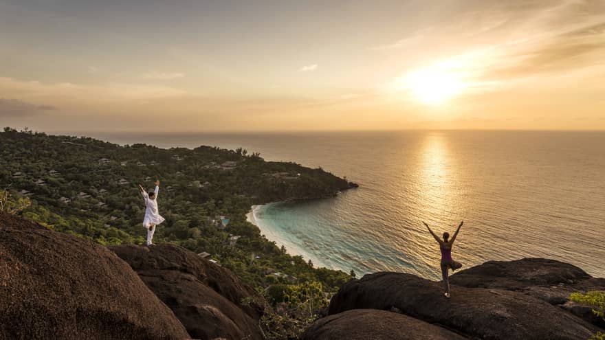 Woman and man stand apart on large boulders overlooking canopy of trees, ocean at sunrise 