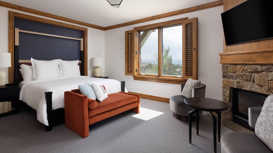 Luxury resort bedroom with a king bed, burnt orange chaise, grey armchairs, stone fireplace, TV and window with wooden shutters