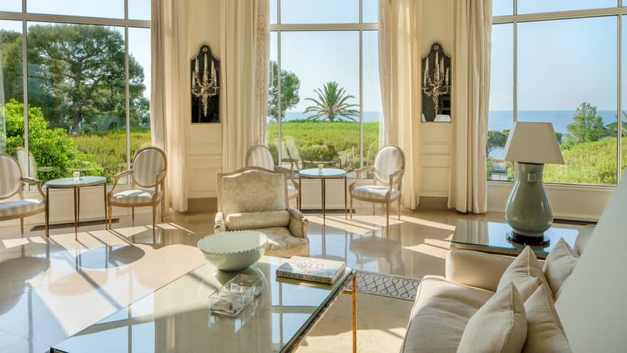 All-white salon with couches and dining chairs with tall rounded windows, lawn and sea view