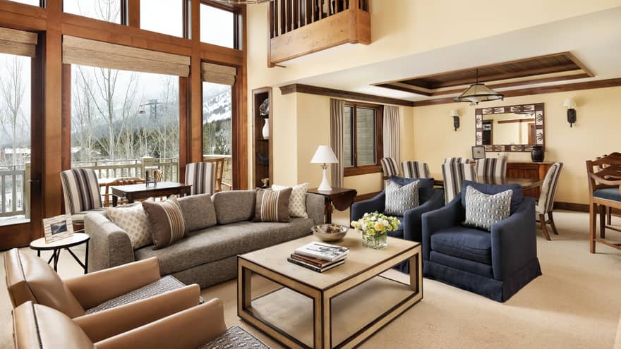 Spacious resort living area with a grey sofa, navy armchairs and large windows offering snowy mountain views, connected to a dining area with striped chairs