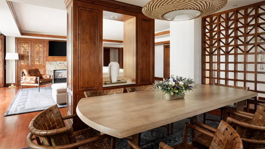 Luxury suite dining area with a large wooden table, woven chairs and a floral centrepiece, with an open view into a cozy lounge with a fireplace