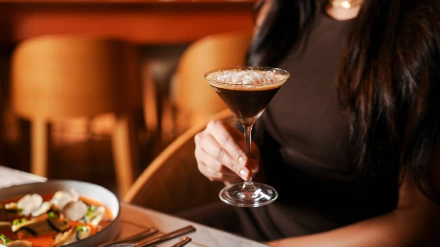 Person with waist-length dark hair and wearing a brown sleeveless dress sits a table in a restaurant holding a martini