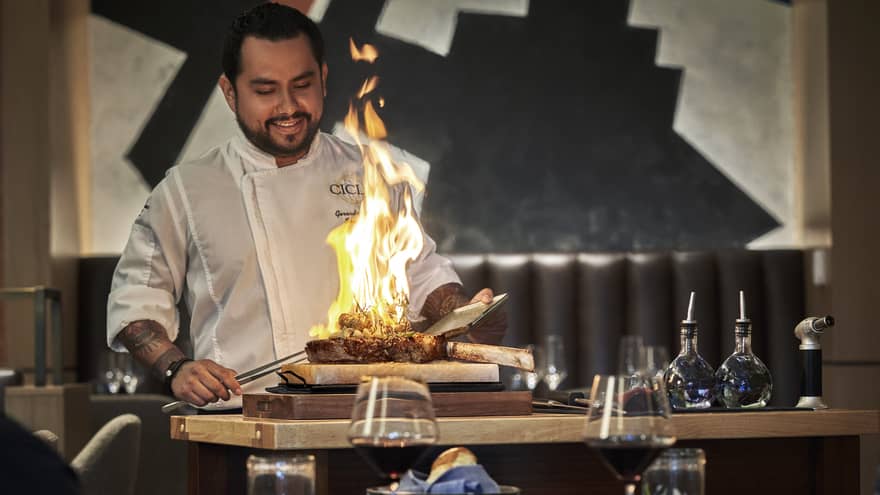 A male chef cooking a large steak with fire in front of people drinking wine.