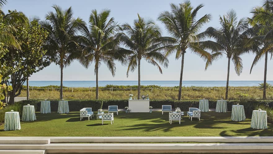 Blue outdoor sofas surround white bar, cocktail tables with long tablecloths on grass surrounded by tall palm trees with an ocean view