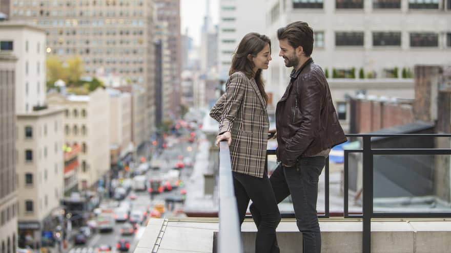Man and woman wearing autumn coats lean in close against balcony overlooking New York City