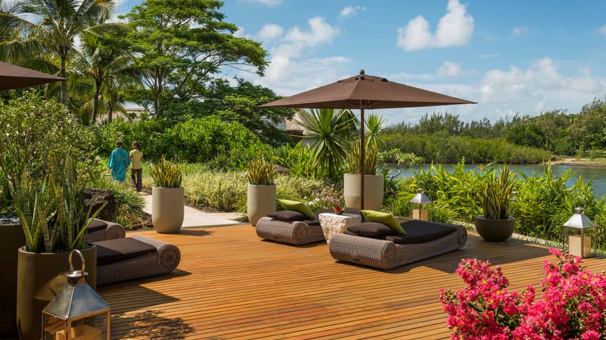 Woman in silk robe and hotel staff walk on path from wicker lounge beds on relaxation patio