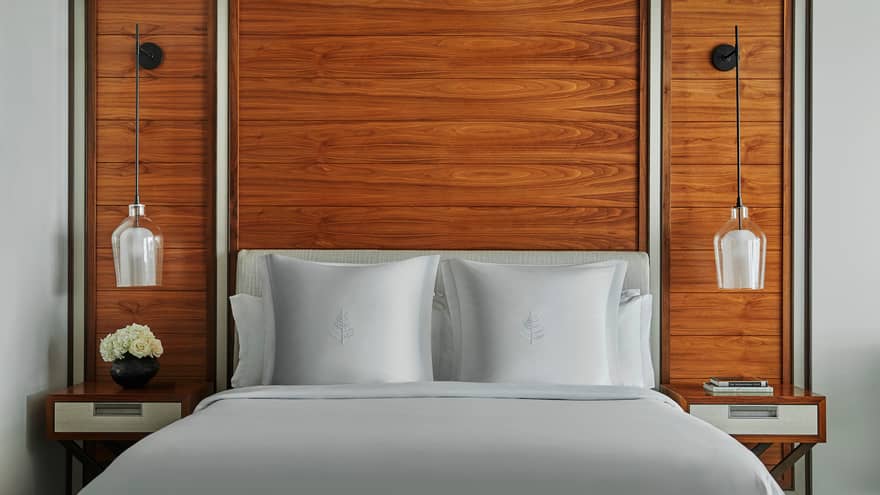 Top of hotel bed, pillows with white linens against wood accent walls, hanging lamps on each side