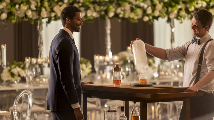 A groom talking to a bartender while the bartender makes a drink, there are flowers hanging in the background.
