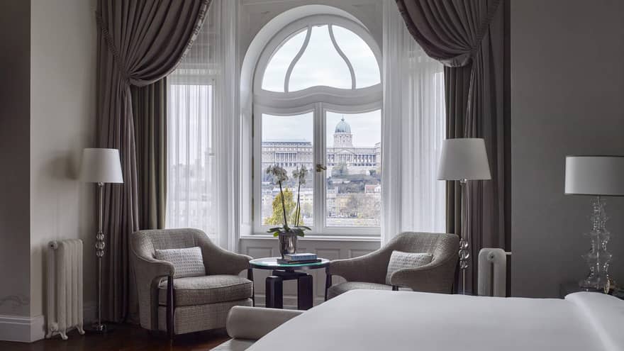 Buda Castle Presidential Suite is furnished with a two sitting chairs next to a curved window, lamps, a large bed and gray curtains. 