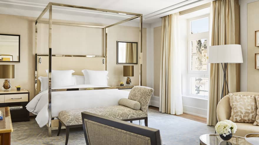 Luxury hotel bedroom with a modern four-poster bed, beige sofa, glass coffee table and large window with city views, decorated in neutral tones