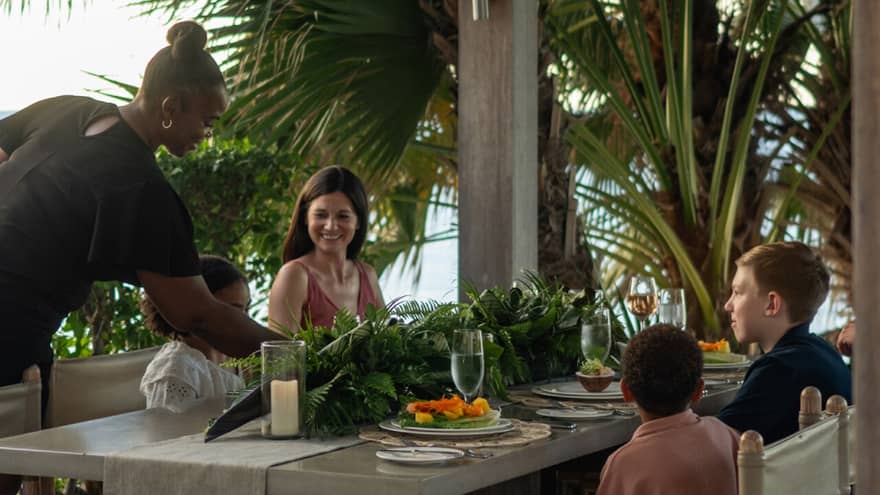 A family smiles as they enjoy a meal at an outdoor dining table surrounded by palm trees