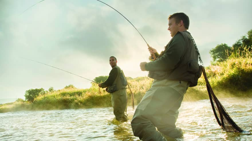 Two people fly fishing in a river, standing knee-deep in the water, surrounded by mist and greenery.