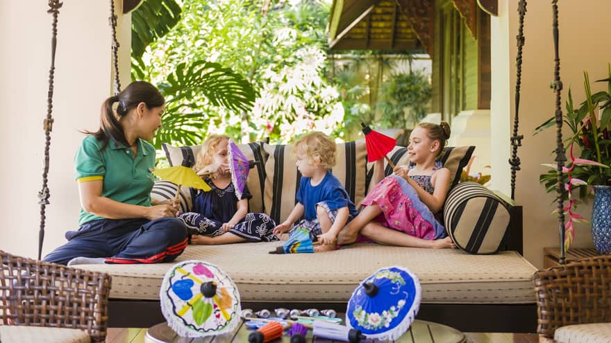 Hotel staff relaxes with children, hold colourful paper umbrellas on large hanging patio bed 
