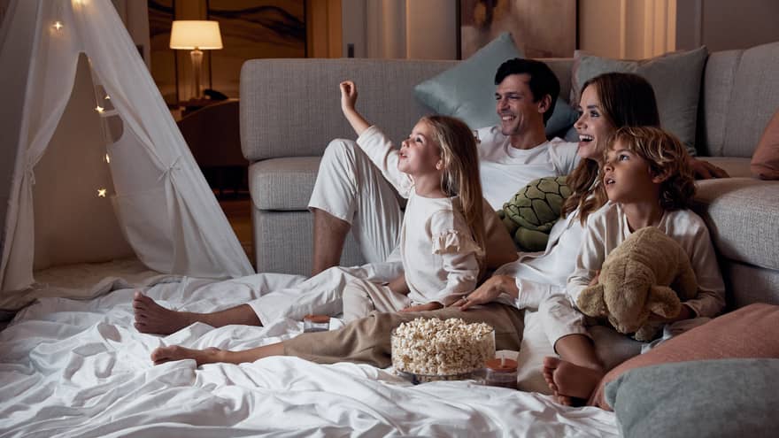 A family relaxing on blankets beside a sofa; the children hold stuffed toys and there's a bowl of popcorn between them.