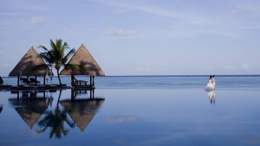 Bride and groom walk along patio infinity pool on ocean, appearing to walk on water