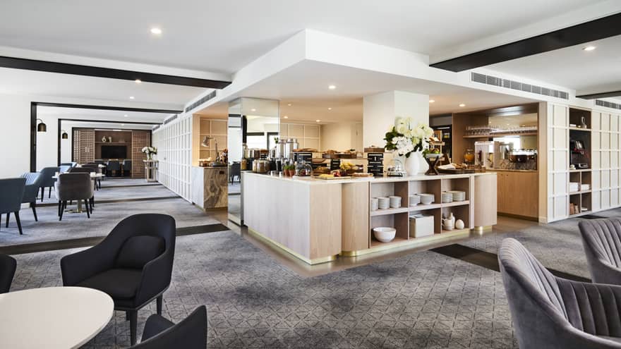 A modern hotel lounge with plush grey chairs and carpet, and a well-equipped kitchen set up with buffet foods and coffees.
