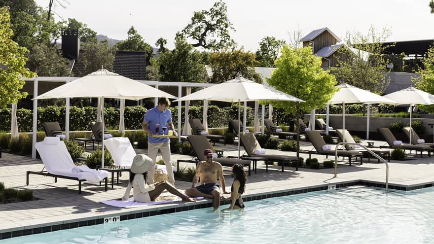 A person bringing a tray of beverages to three guests near the pool