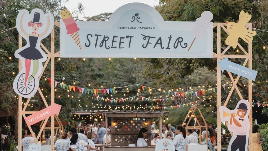 A colorful archway with a sign that reads "Street Fair" serves as an entrance to a street decorated with sting lights, multi-color pendant banners and booths