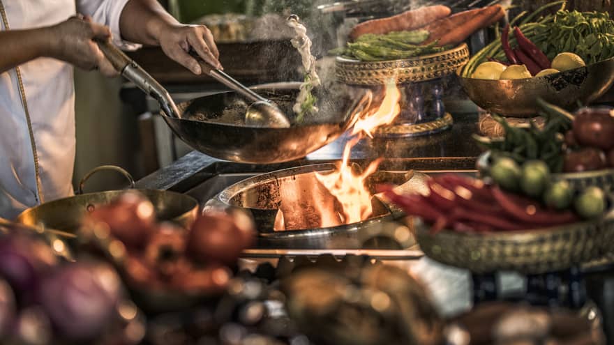 Close-up of chef stirring meals in copper pans over stove, flames 