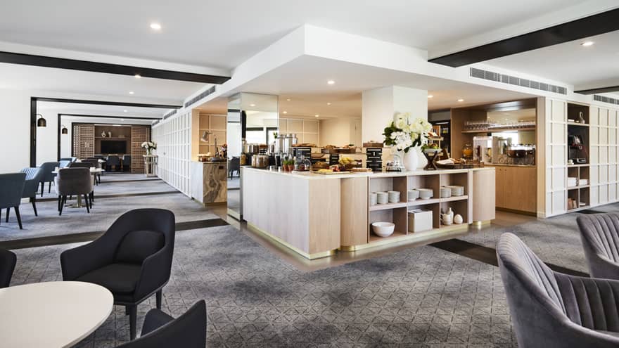 A modern hotel lounge with plush grey chairs and carpet, and a well-equipped kitchen set up with buffet foods and coffees.