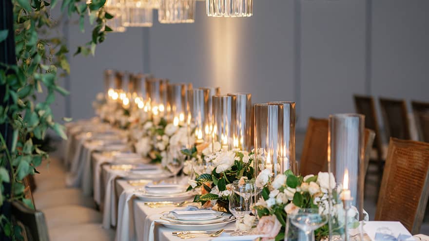 Long dining table elegantly set with candles, white floral arrangements, and fine tableware, creating a sophisticated ambiance.,Long dining table elegantly set with candles, white floral arrangements, and fine tableware, creating a sophisticated ambiance.