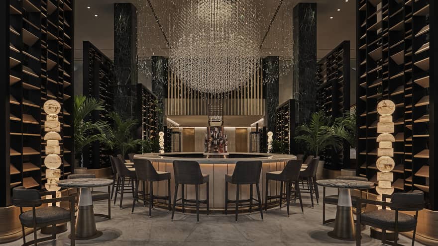 A bar area surrounded by chairs and small tables and a large chandelier above the bar.