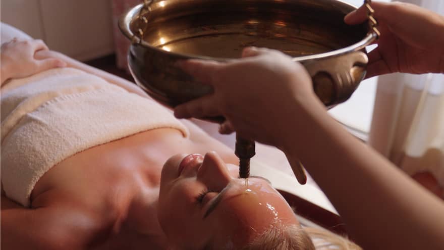 Person lays on massage table covered in a white towel as a spa attendant guides a hanging brass bowl filled with oil over the person's face