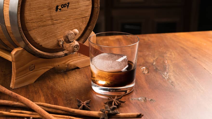 Rock glass with large ice cube, ounce of dark rum in front of small wood barrel on bar 
