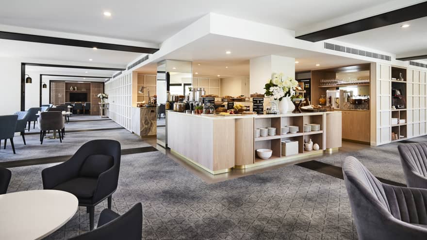 A modern hotel lounge with plush grey chairs and carpet, and a well-equipped kitchen set up with buffet foods and coffees.