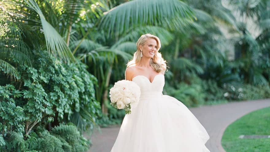 A bride in a white ballgown style wedding dress holds a white bouquet and is surrounded by green, lush vegetation