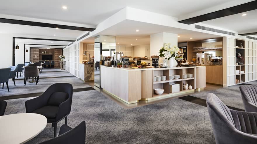 A modern hotel lounge with plush grey chairs and carpet, and a well-equipped kitchen set up with buffet foods and coffees.