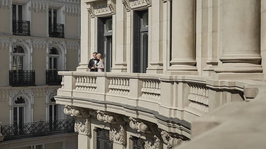 A couple in formal attire stands on a grand balcony of an elegant building, sharing a moment while holding champagne glasses, with ornate architecture in the background.