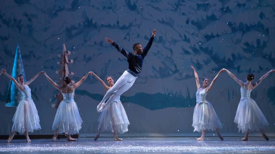 Ballerinas on stage in long ice-blue tutus dance before a snowy painted backdrop as a dancer wearing a leotard leaps grandly.