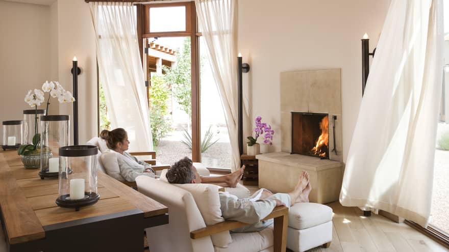 Man, woman in white bathrobes relax on spa armchairs by fireplace, white curtains