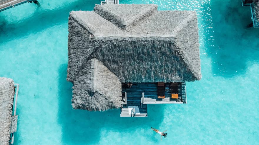Aerial view of person swimming away from overwater bungalow in turquoise lagoon