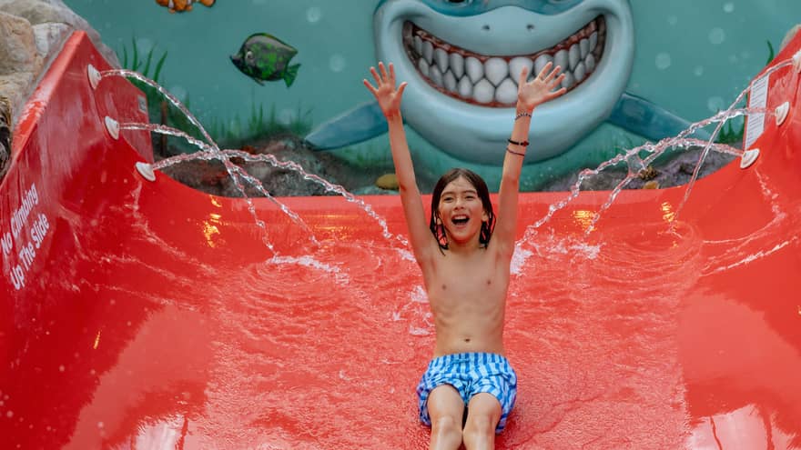 Child wearing blue-and-white striped swim trunks slides with their hands up down a bright red water slide with a mural of a smiling shark in the background