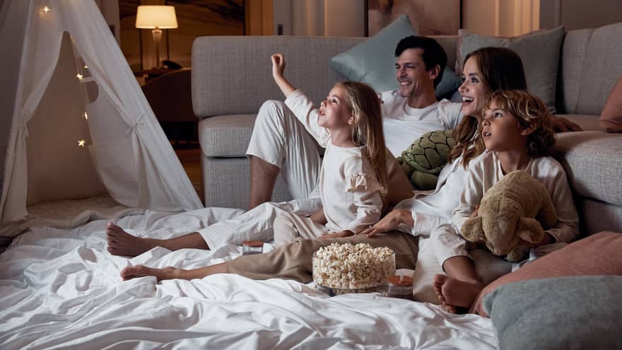 A family relaxing on blankets beside a sofa; the children hold stuffed toys and there's a bowl of popcorn between them.