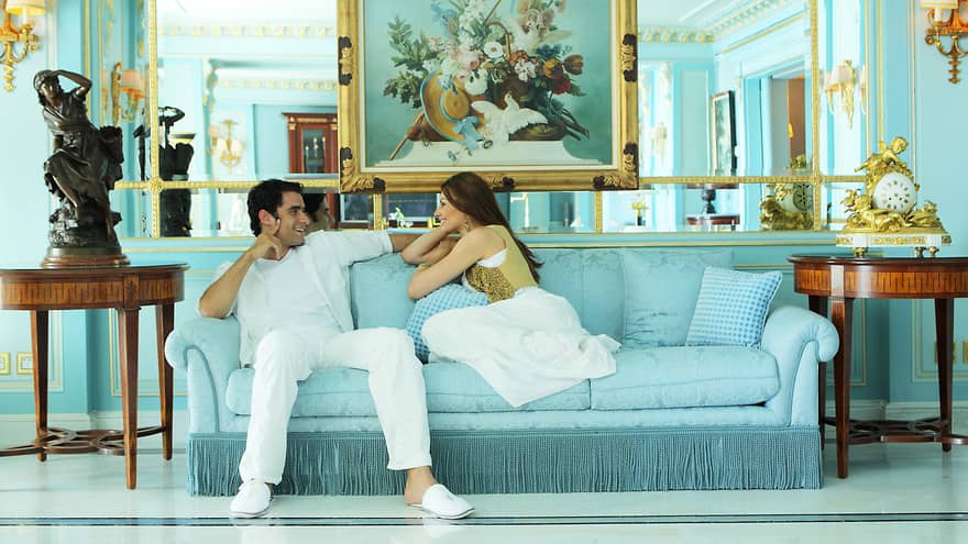 Man and woman smile, relax on blue sofa in Presidential Suite below large mirror, painting, beside statues 