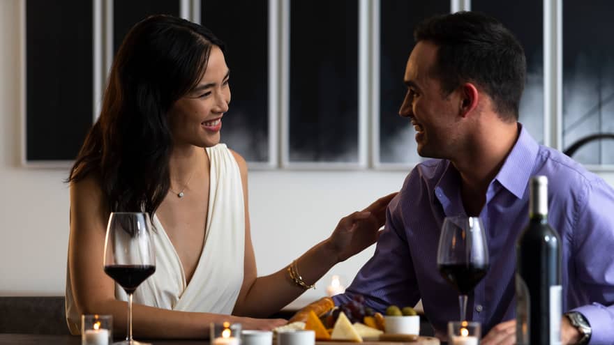 A couple exchanges smiles as they enjoy a cheese platter and a bottle of wine.