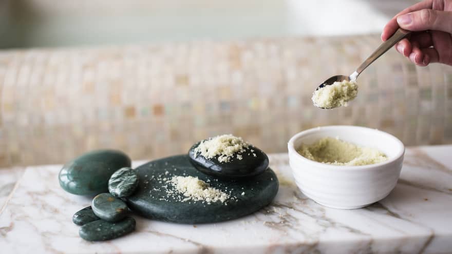 Silver spoon scoops spa salts out of small white bowl, black lava rocks piled nearby on white marble counter