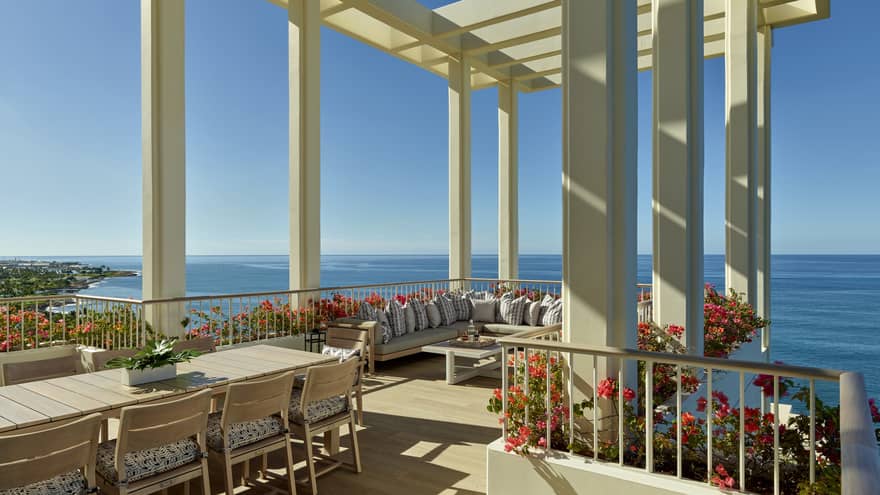 A spacious terrace with a pergola overlooking the ocean, featuring a large dining table, comfortable seating and vibrant flower arrangements under a clear blue sky