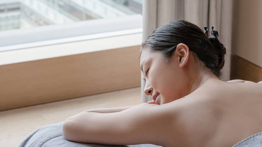 Person relaxing on massage table by a window with eyes closed, enjoying a peaceful moment
